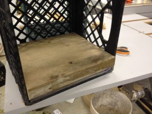 A milk crate is the perfect dimension for sandpaper sheets, and this concrete-form 2x12 cutoff is just the right amount of weight to hold the paper flat.